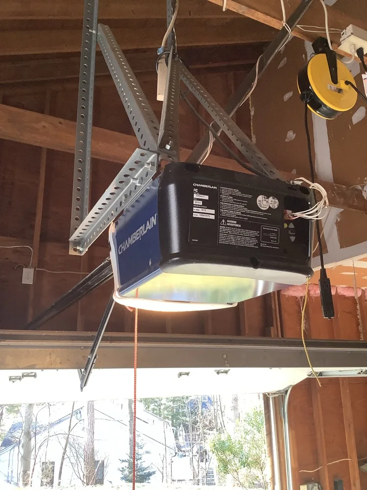 A Chamberlain opener head hung from punched angle-iron under bare joists, its light lens lit and its release cord dangling, with the open bay and leafless woods visible below.