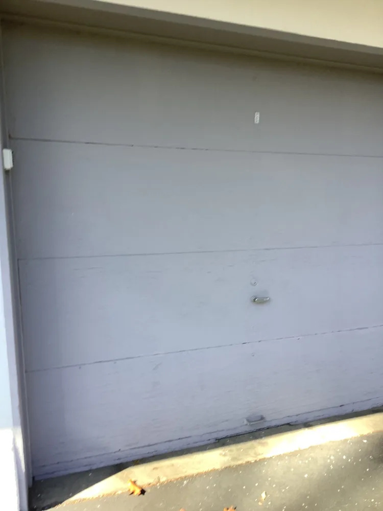 A grey-lilac painted flush garage door photographed close on, its lift handle at mid height and fallen brown leaves gathered along the threshold.