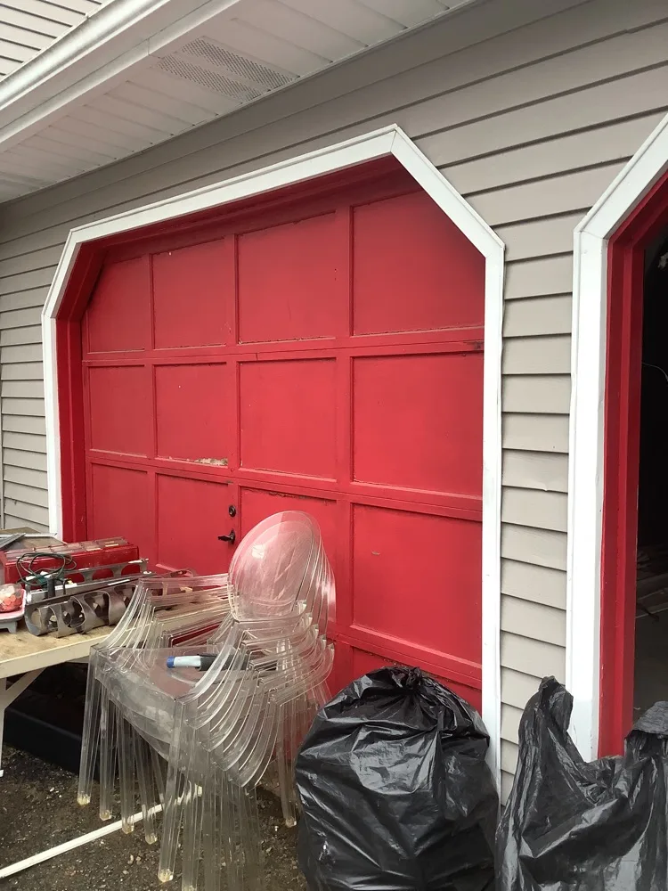 A red painted wood carriage door with a shallow arched head and white surround, set into grey clapboard siding beside a second matching opening.