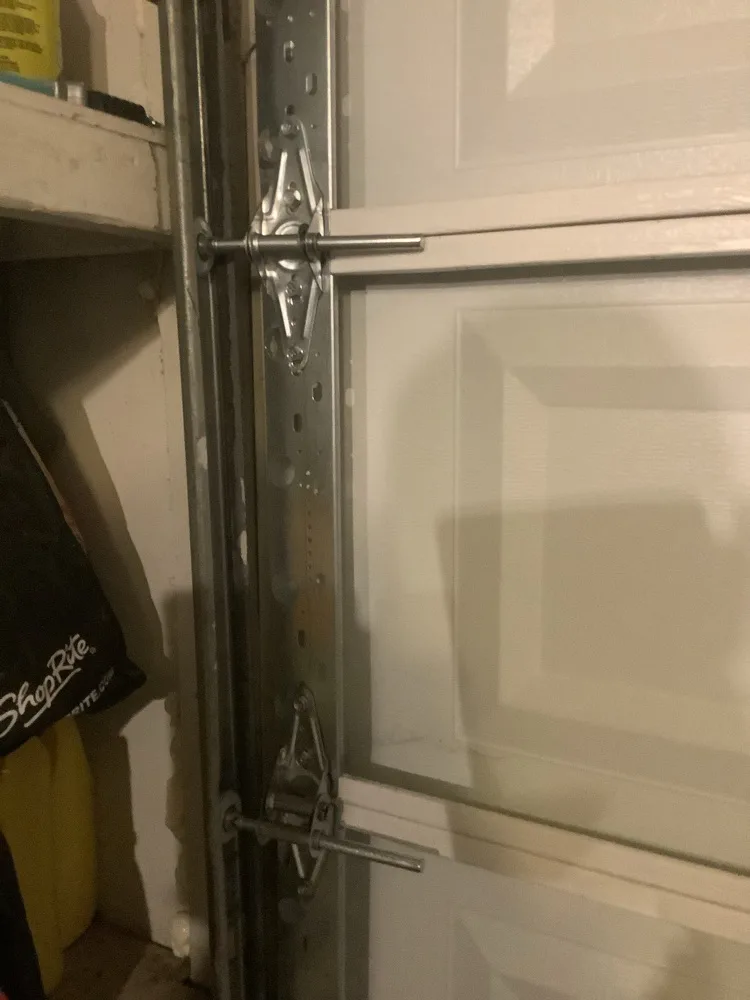 The vertical track of a garage door photographed from inside, two hinges and their roller stems bolted through the sections and seated in the track, with garage shelving to one side.