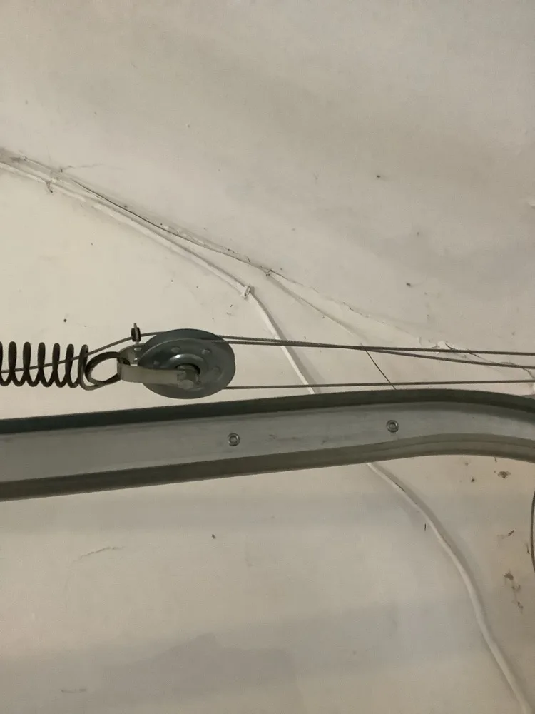 An extension spring hooked to a pulley above the horizontal track of a garage door, the doubled lift cable running away from it along a white ceiling.
