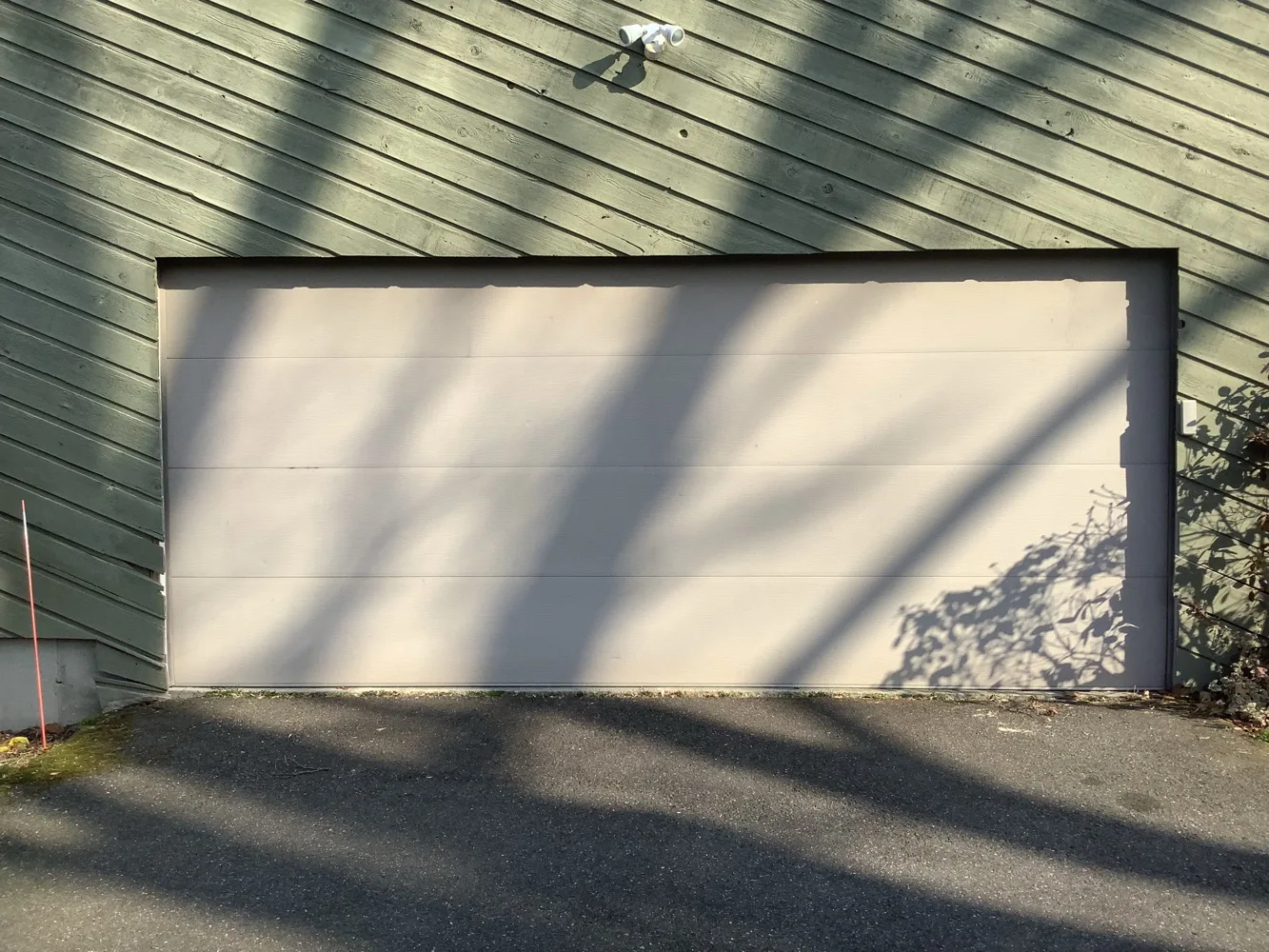 A wide flush-panel garage door in pale sage board siding, the shadows of bare tree branches falling across the door and the asphalt apron in front of it.