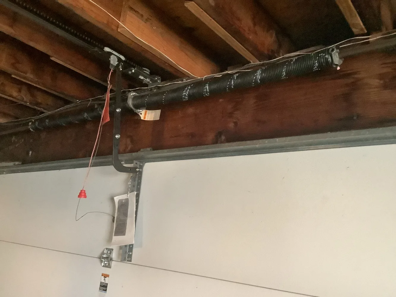 A black torsion spring wound onto its shaft along the header above a garage door, bolted through a centre bearing plate, with the opener rail and its red emergency release cord alongside and rough joists overhead.