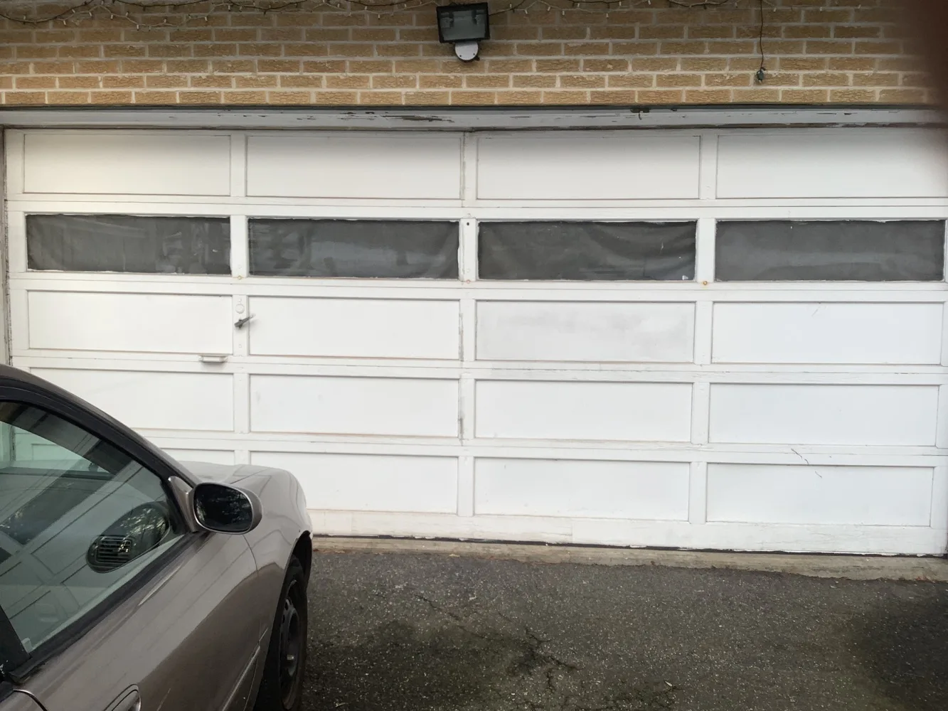 A white long-panel garage door with a band of dark windows set into tan brick, a saloon car parked close in front of it on cracked asphalt.